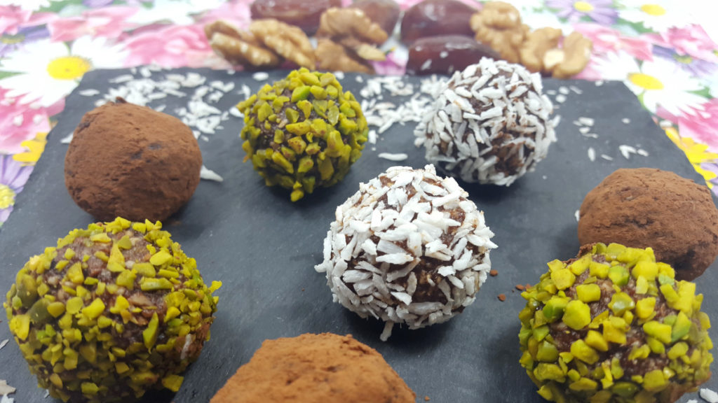 Bliss Balls Energy Balls aus Datteln mit Kokosraspeln, Pistazienkernen, Kakaopulver ummantelt. Auf einem schwarzen Brett auf Blumenuntergrund.