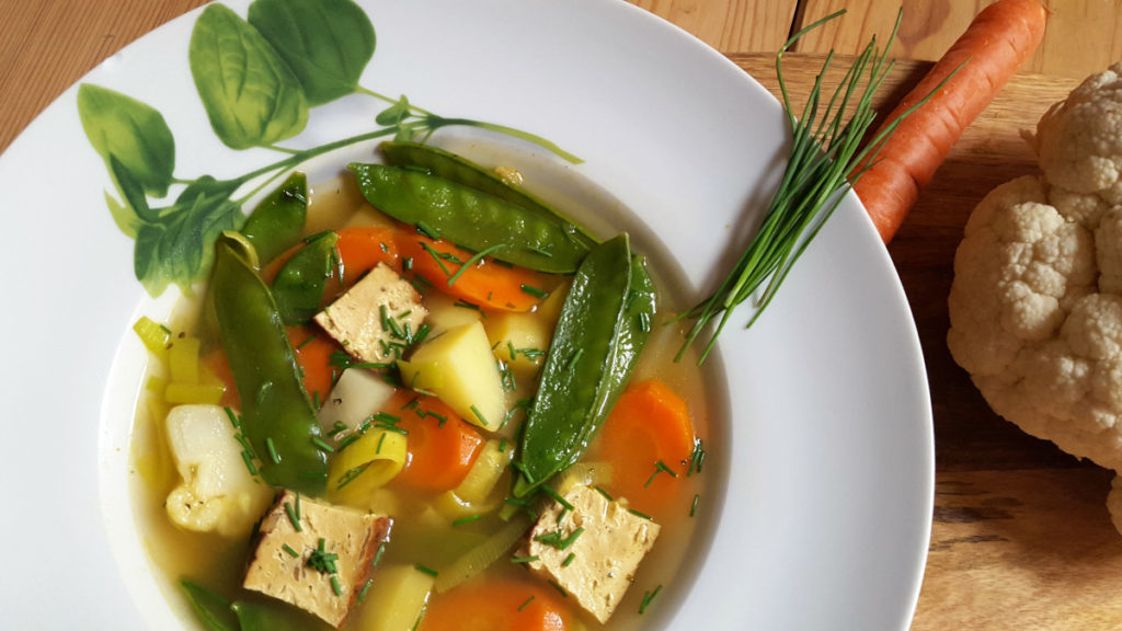 Gemüsesuppe mit Möhren, Kohlrabi, Zuckerschoten, Lauch, Kartoffeln, Räuchertofu auf weißem Teller mit Basilikumdeko, Schnittlauch auf dem Rand, Möhre und Blumenkohl angelegt